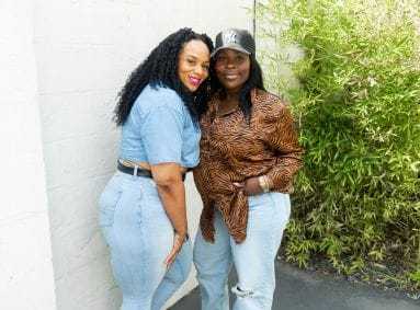 Two women smiling and posing together in casual outfits outdoors near a bamboo plant.