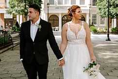 Bride and groom holding hands, standing outdoors in wedding attire, facing opposite directions.