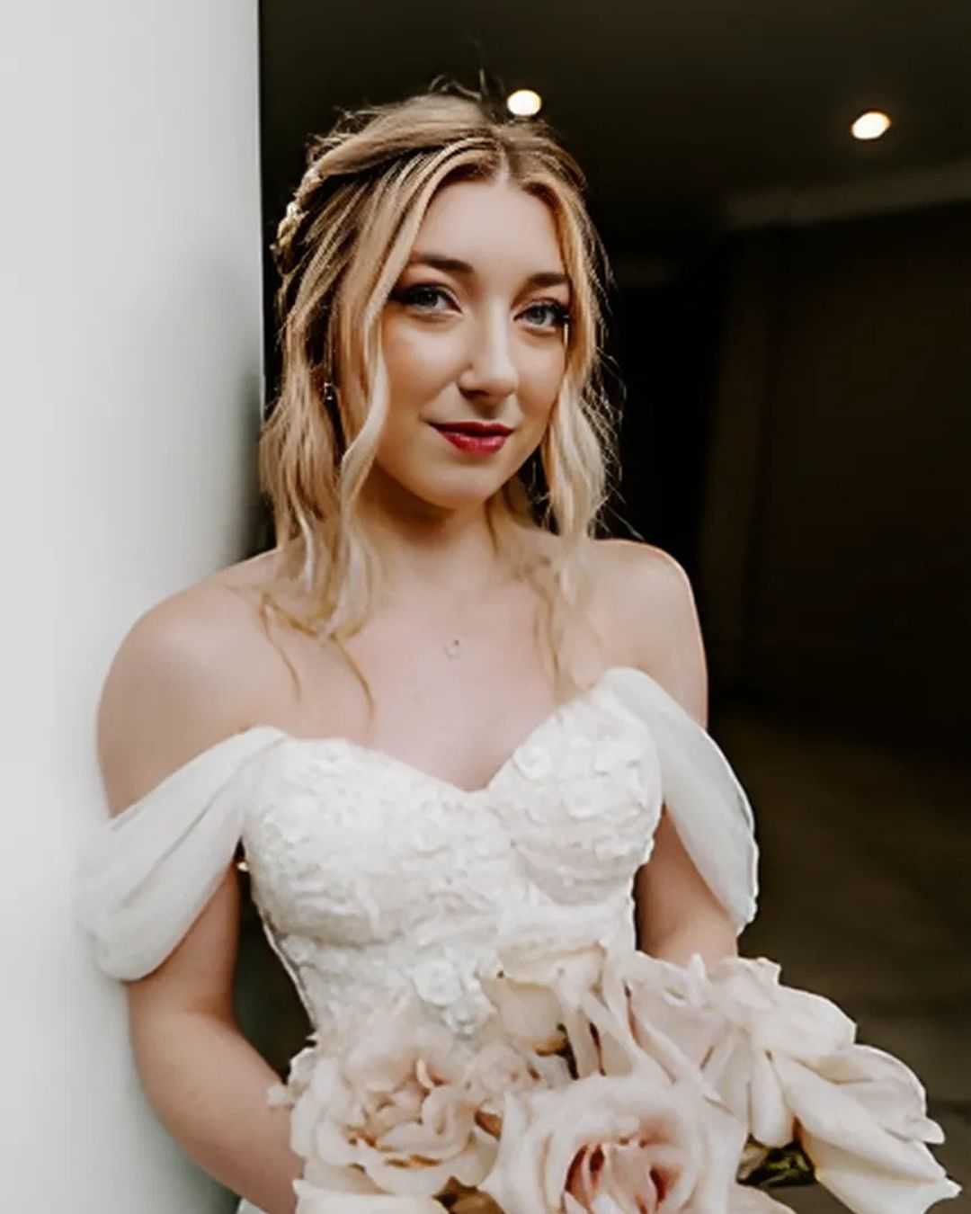 Bride in an off-shoulder white gown holding a bouquet of pale pink roses, smiling indoors.