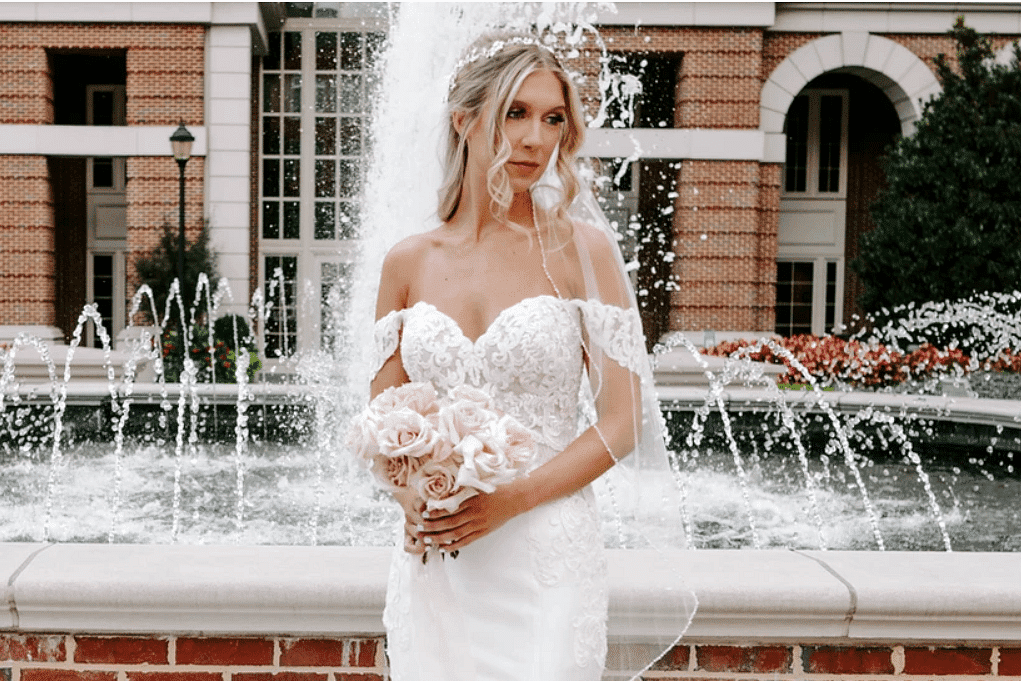 Bride in white dress holding bouquet, standing in front of a fountain with brick building background.