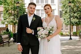 Bride and groom standing outdoors in a courtyard, holding a bouquet and looking at the camera.