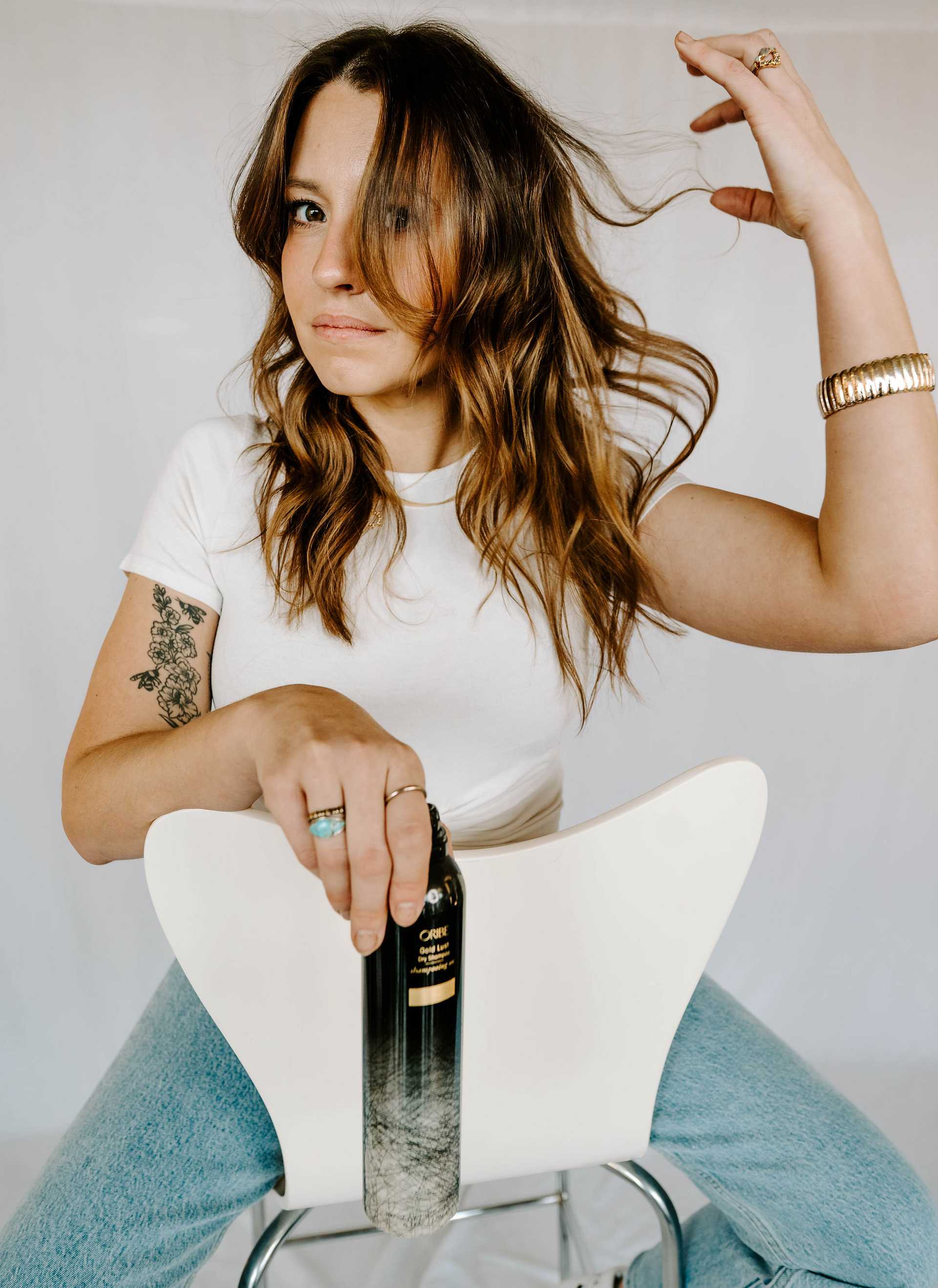 Woman sitting backward on chair, holding bottle. Touching hair with one hand, wearing white shirt.